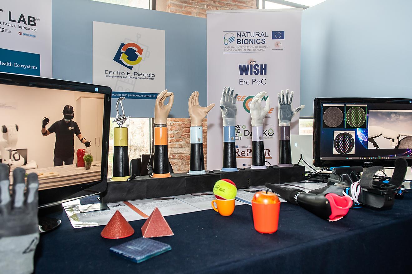 Mani robotiche e protesiche esposte su un tavolo con oggetti colorati per i test di presa, in uno stand del Festival.