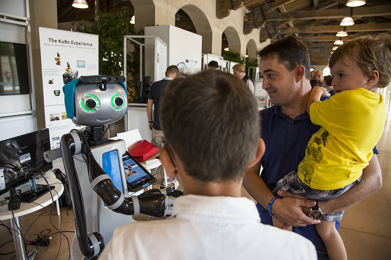 Un padre con un bambino in braccio osserva un piccolo robot dagli occhi luminosi in uno stand espositivo del Festival della Robotica.