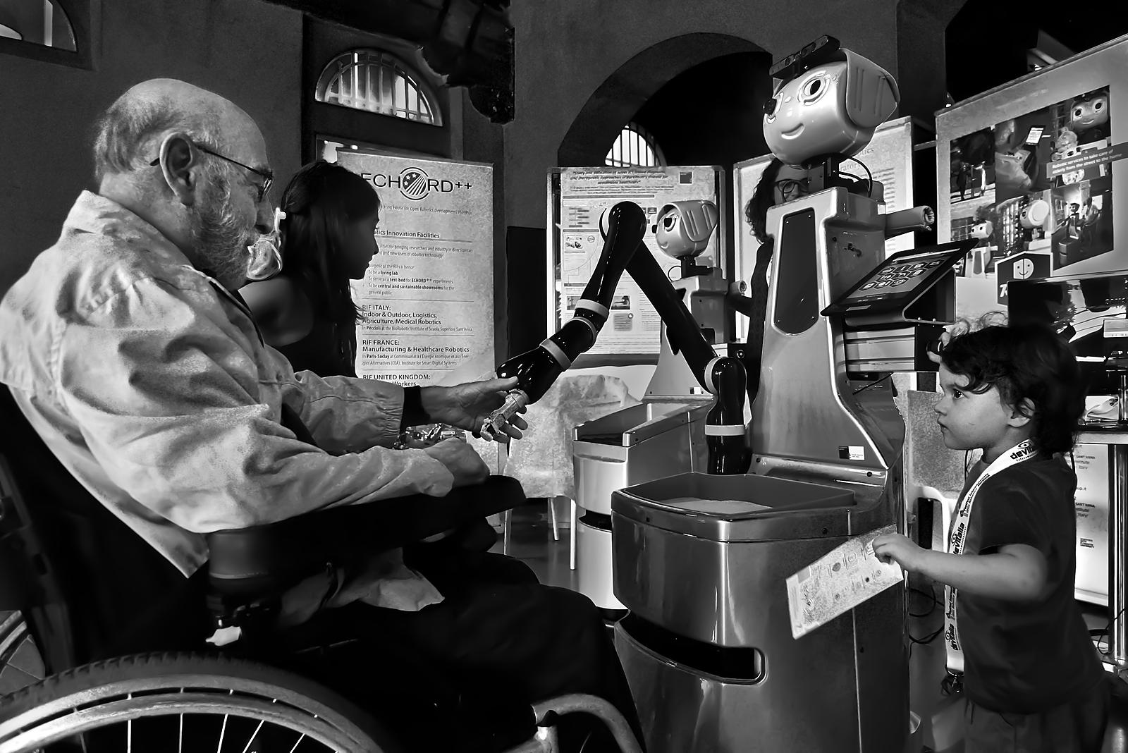 Un braccio robotico di servizio aiuta una persona anziana in carrozzina mentre una bambina osserva: robotica assistiva per l'autonomia quotidiana.